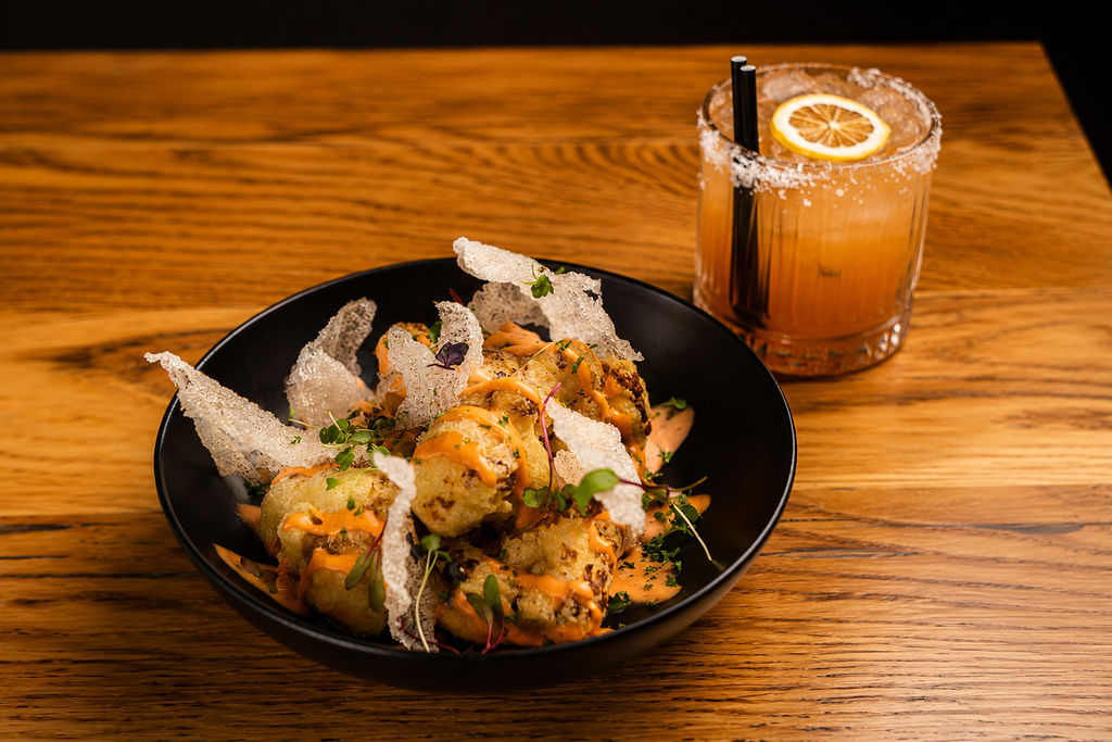 A bowl of crisp fried cauliflower with chilli sauce, crackers and micro herbs, with a cocktail alongside