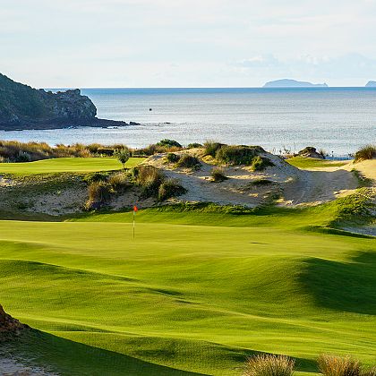 A green and flag on the links, dune grasses and the Pacific beyond, with a headland to one side and an island on the horizon